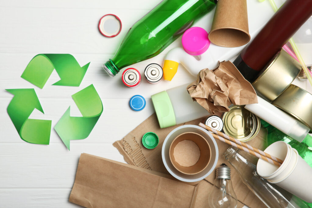 Collection of recyclable materials including a green plastic bottle, crumpled brown paper and cardboard rolls, metal cans with visible lids, plastic caps in various colors, an empty adhesive tape roll, and a light bulb scattered around a prominent green recycling symbol.