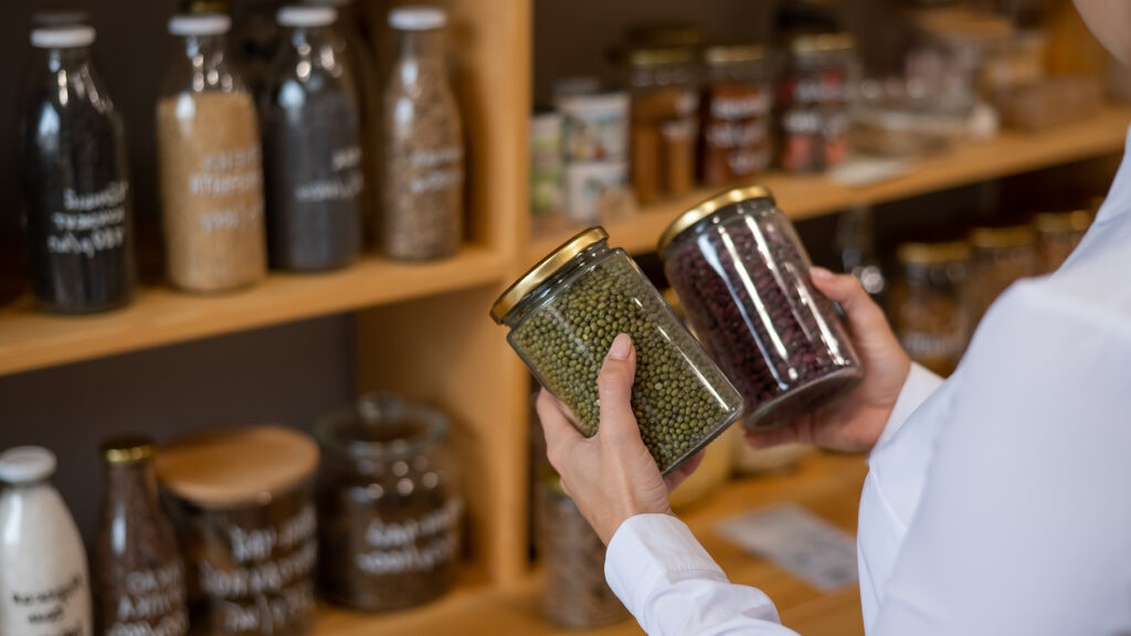 A person's hands holding two clear glass jars filled with green mung beans and red kidney beans, with blurred shelves of various labeled jars in the background, showcasing an organized, eco-friendly approach Smart Eco-Storage Solutions.