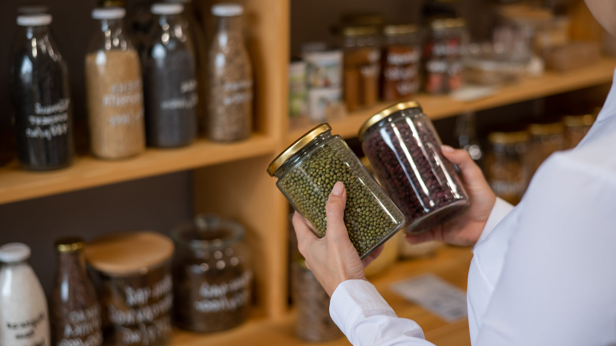 A person's hands holding two clear glass jars filled with green mung beans and red kidney beans, with blurred shelves of various labeled jars in the background, showcasing an organized, eco-friendly approach Smart Eco-Storage Solutions.